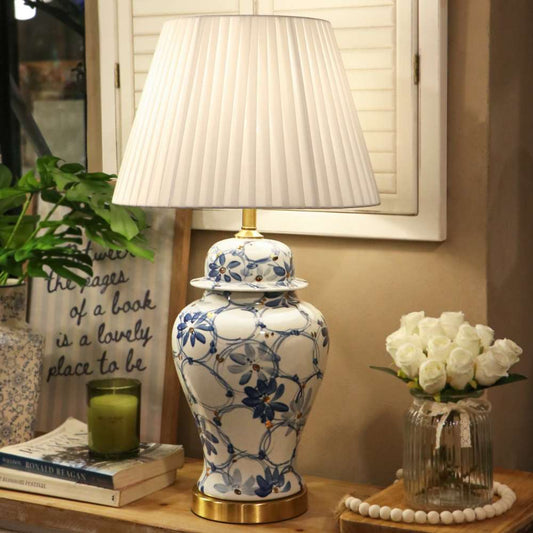 A decorative table lamp with a white pleated drum shade and a ceramic base featuring a blue floral and vine pattern, set on a wooden table in a styled room next to a candle, books, and a vase of white roses.