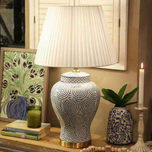 A decorative table lamp with a white pleated drum shade and a ceramic jar-shaped base featuring a blue and white spiral pattern, set on a wooden surface in a styled interior scene.