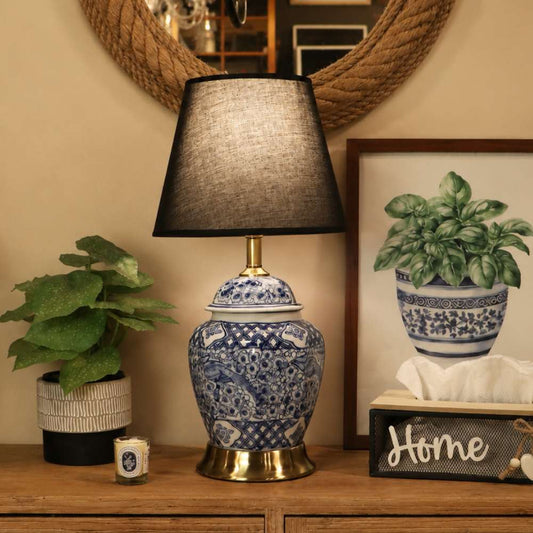 A decorative table lamp with a blue and white patterned ceramic jar base and a black fabric drum shade, placed on a wooden console in a styled room setting.