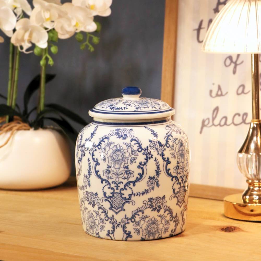 A blue and white patterned ceramic lidded jar sits on a wooden tabletop in a styled interior setting, with a potted white orchid on the left and a lit table lamp on the right.