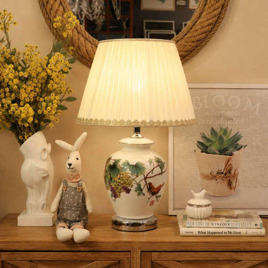 A decorative table lamp with a pleated cream shade and a white ceramic base featuring painted fruit and bird motifs, styled on a wooden cabinet beside a rabbit figurine, flowers, and framed wall art.