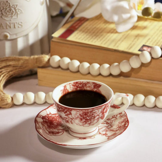 A white ceramic cup and saucer set decorated with a red floral toile pattern, filled with black coffee, styled on a table with books, beads, and a decorative vase in the background.