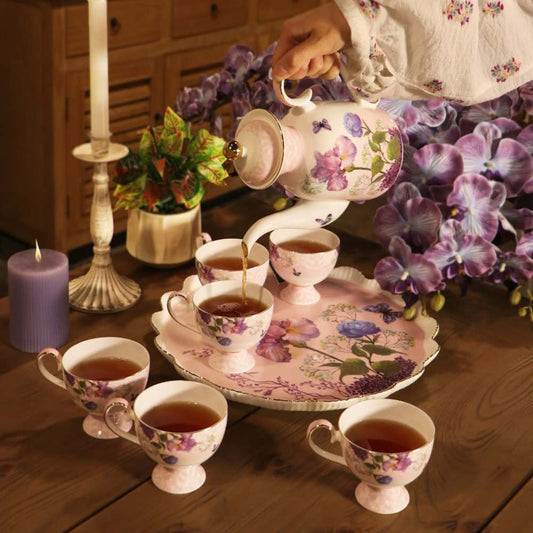 A floral porcelain tea set arranged on a wooden table, with a hand pouring tea from a matching teapot into six teacups on a tray. The set includes a teapot, six teacups, and a serving tray, decorated with purple and blue flowers.