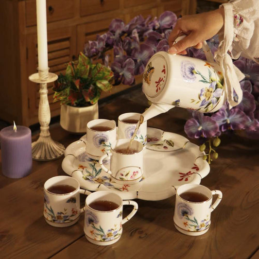 A ceramic tea set with a teapot, matching cups, and a serving tray decorated with floral patterns, arranged on a wooden table with a hand pouring tea. The scene includes candles, a small plant, and purple flowers in the background.