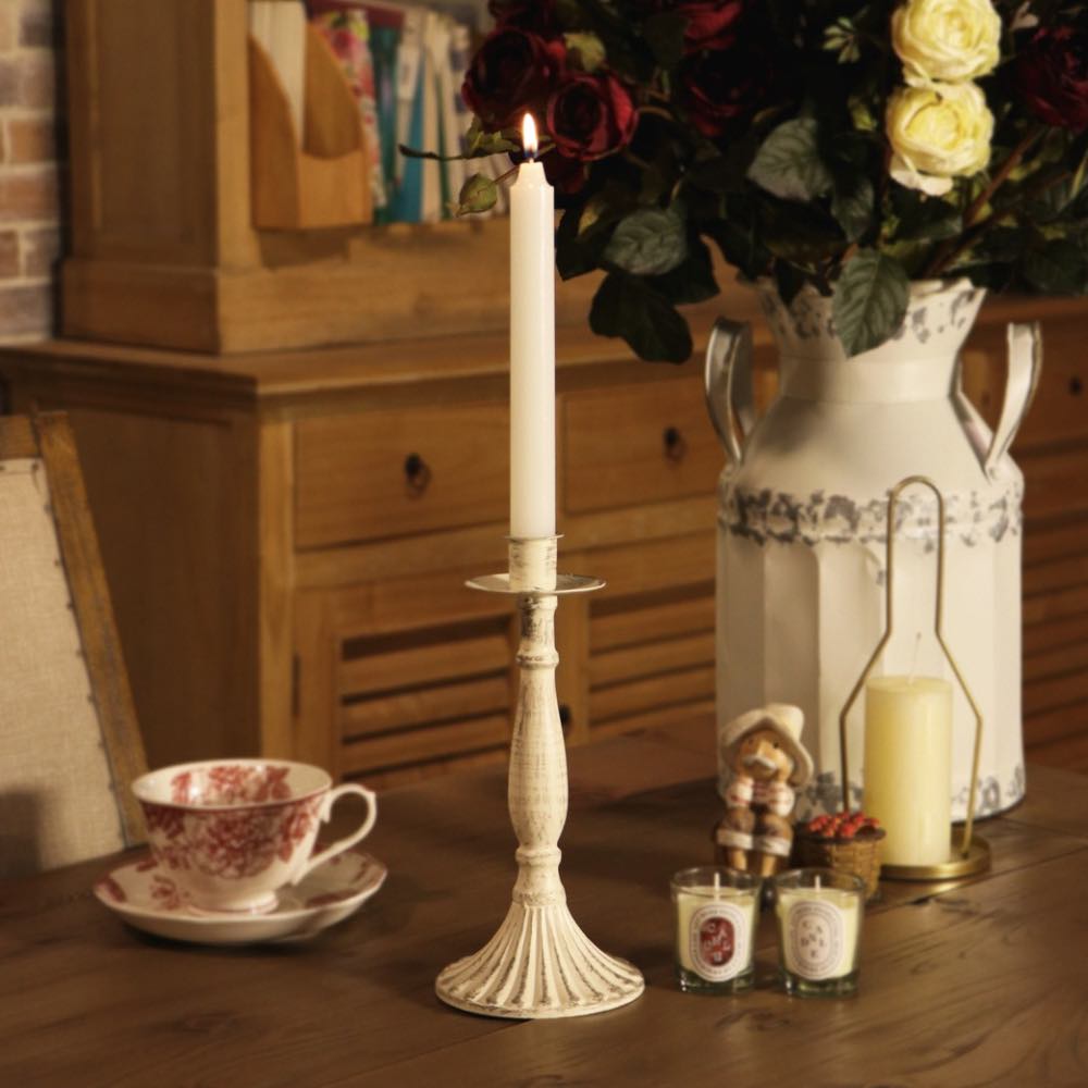 A tall white taper candle in a ribbed cream-colored metal holder sits on a wooden table, styled with a matching teacup, small candles, a decorative figurine, and a large white pitcher filled with red and yellow roses.