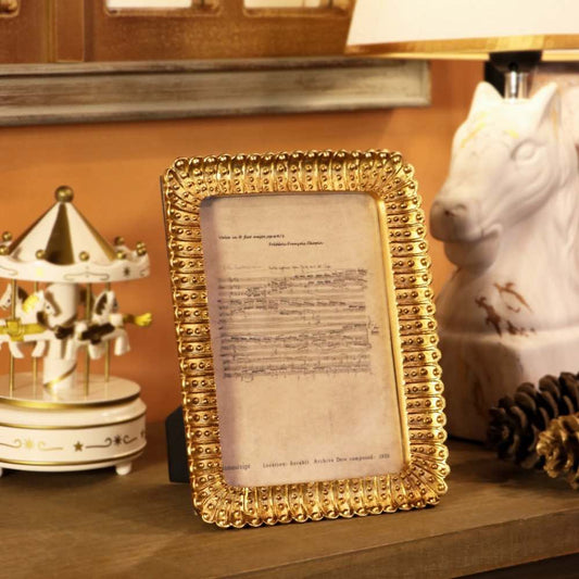 A rectangular picture frame with an ornate textured gold border, holding a vintage sheet music print, displayed on a wooden shelf next to a white carousel figurine and a white horse head lamp.