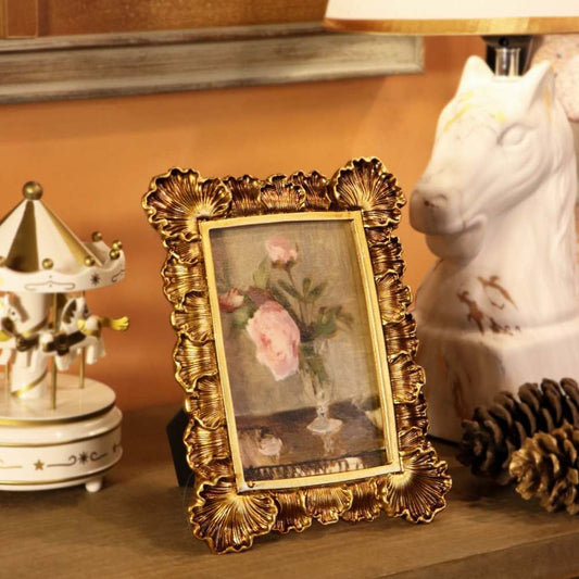 An ornate gold picture frame with a vintage floral painting, styled on a wooden shelf next to a white ceramic horse lamp and a miniature carousel toy.