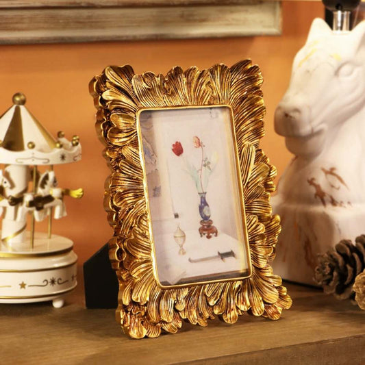 An ornate gold picture frame with a leaf-like scalloped border, holding a small floral photo, displayed on a wooden surface next to a white carousel and a white horse figurine.