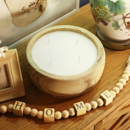 A white four-wick candle in a round light wood bowl, styled on a wooden table with a beaded 'HOME' garland, a small picture frame, and a ceramic vase in the background.