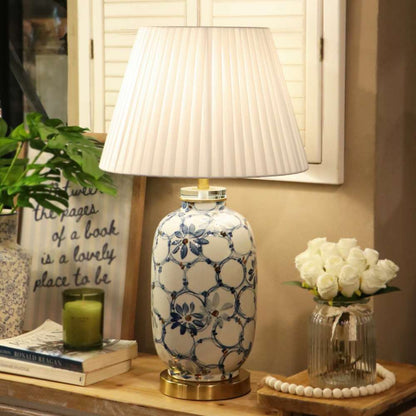A decorative table lamp with a white pleated shade and a ceramic base featuring a blue floral and geometric pattern, styled on a wooden shelf next to a candle, books, and white roses.