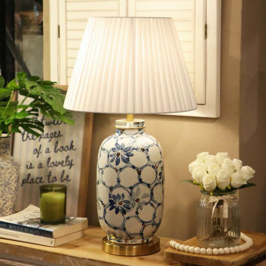 A decorative table lamp with a white pleated shade and a ceramic base featuring a blue floral and geometric pattern, styled on a wooden shelf next to a candle, books, and white roses.
