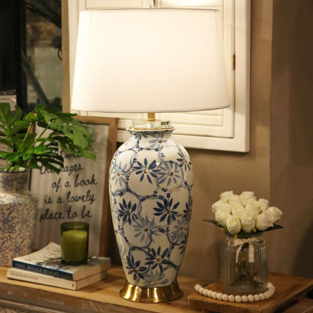 A table lamp with a white fabric drum shade and a ceramic base decorated in a blue and white floral pattern with gold-toned trim, styled on a wooden console table in a home interior setting.