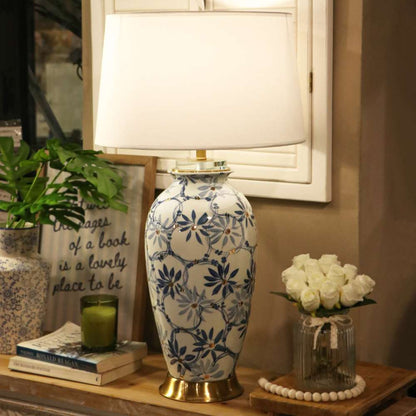 A table lamp with a white fabric drum shade and a ceramic base decorated in a blue and white floral pattern with gold-toned trim, styled on a wooden console table in a home interior setting.