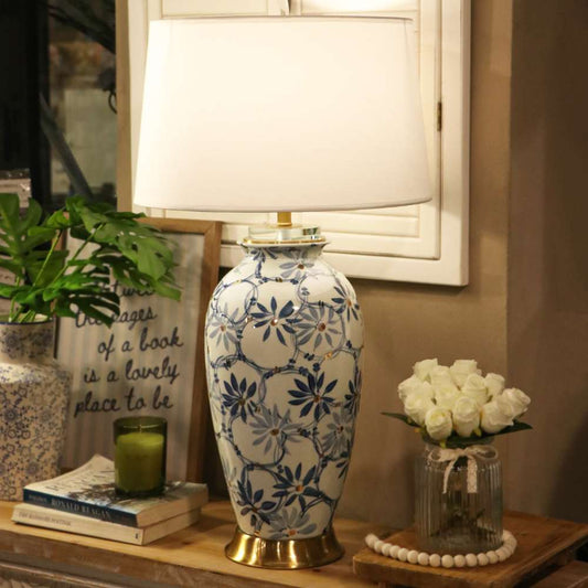 A table lamp with a white fabric drum shade and a ceramic base decorated in a blue and white floral pattern with gold-toned trim, styled on a wooden console table in a home interior setting.
