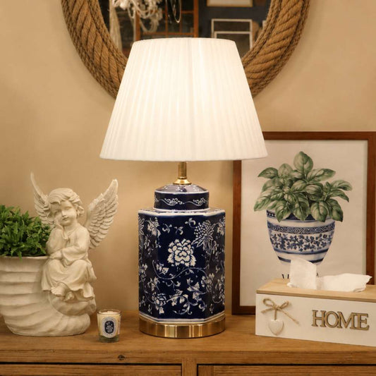 A table lamp with a white pleated drum shade and a blue-and-white floral ceramic base sits on a wooden console table in a styled room setting, alongside a white angel statue, a small candle, a tissue box, and framed wall art.