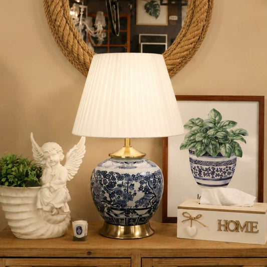 A table lamp with a white pleated drum shade and a blue-and-white porcelain-style ceramic base with brass accents, styled on a wooden cabinet beside a white angel figurine, a tissue box, and framed artwork.