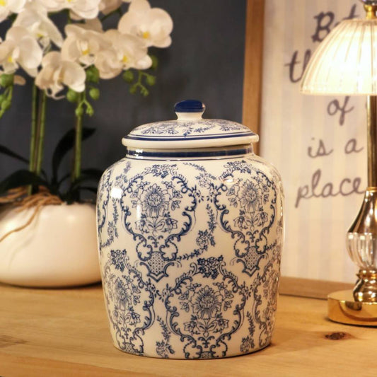 A ceramic storage jar with a matching lid, decorated in a classic blue and white damask pattern, set on a wooden table next to a lit table lamp and white orchids.
