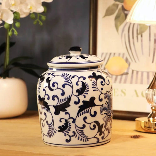 A white ceramic jar with a matching lid, decorated with a dark blue floral and scroll pattern, displayed on a wooden tabletop with a lamp and flowers in the background.