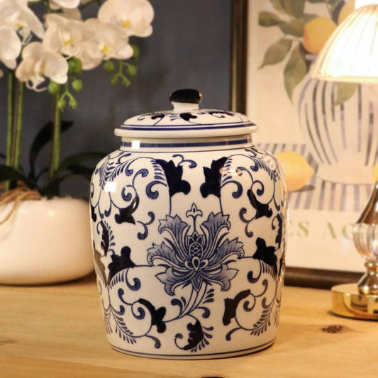 A white ceramic jar with a matching lid, decorated with a blue and gray floral pattern, set on a wooden table in a styled home environment with a lamp and orchids in the background.