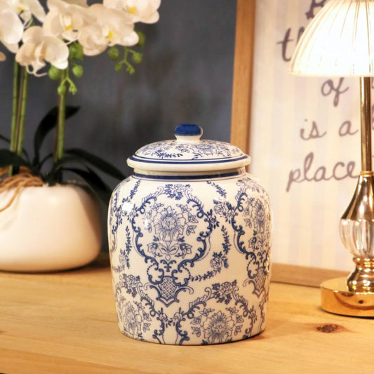 A blue and white patterned ceramic lidded jar sits on a wooden tabletop in a styled interior setting, with a potted white orchid on the left and a lit table lamp on the right.