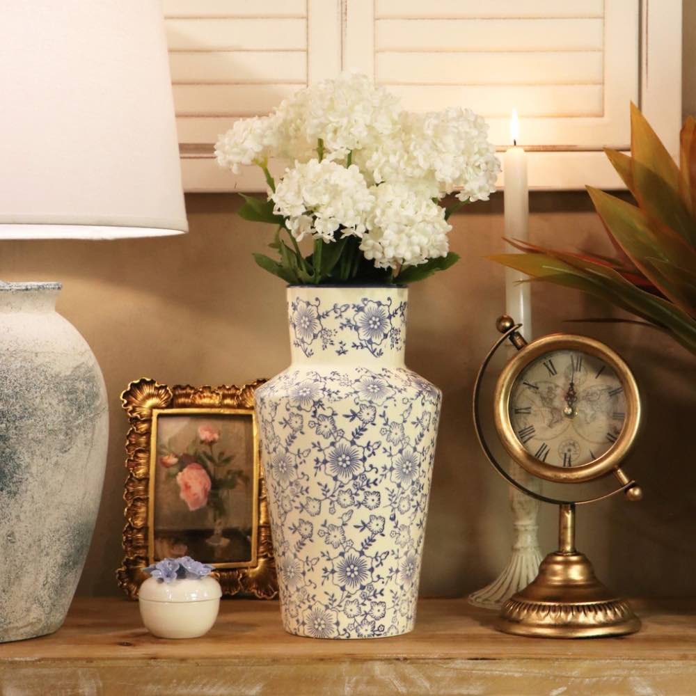 A tall ceramic vase with a blue floral pattern on a cream background, filled with white hydrangea blooms, displayed on a wooden shelf next to a lamp, a small framed picture, a candle, and a brass clock.