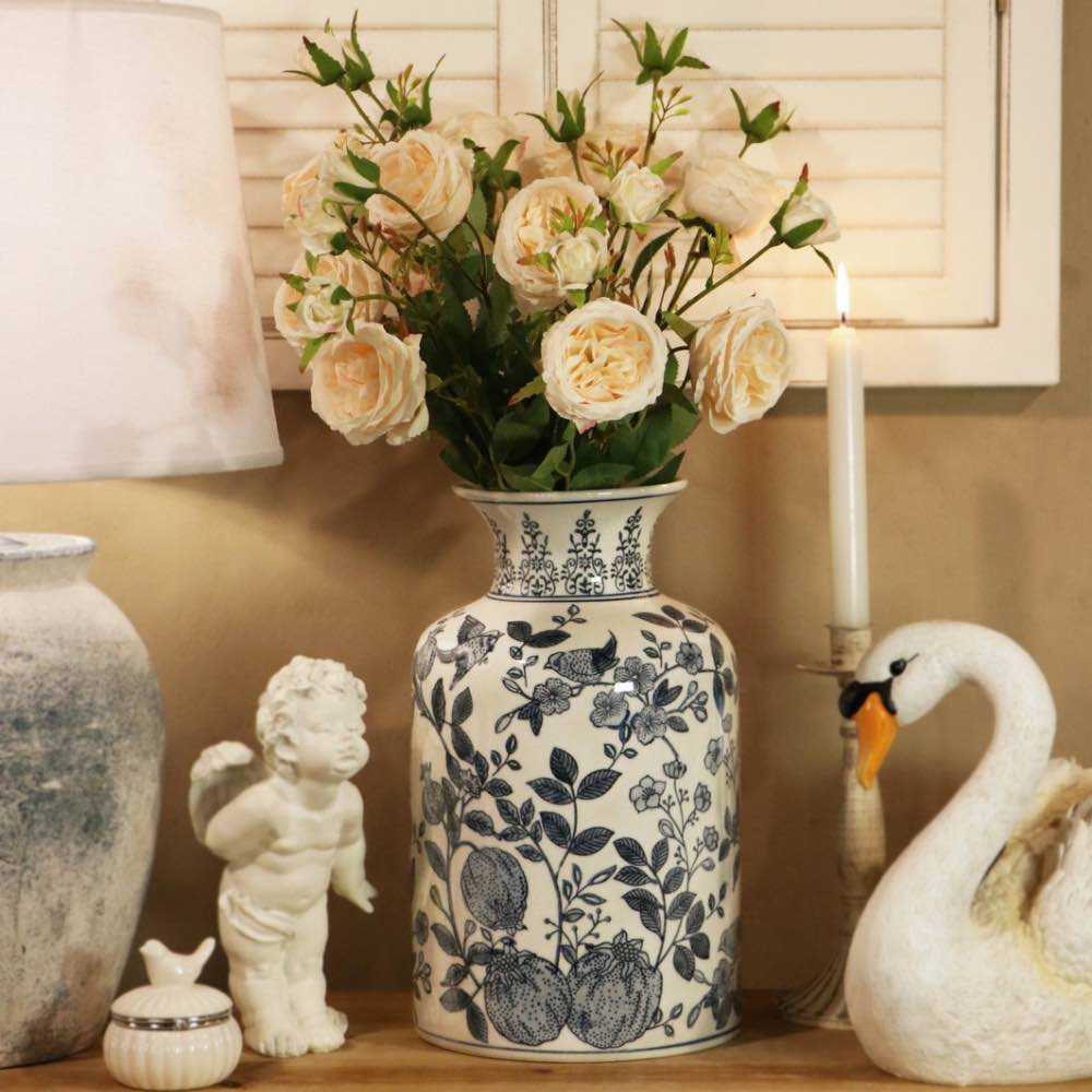 A tall ceramic vase with a blue and white floral pattern, filled with cream-colored faux roses, displayed on a shelf next to a white swan figurine, a small white cherub statue, and a lit candle.