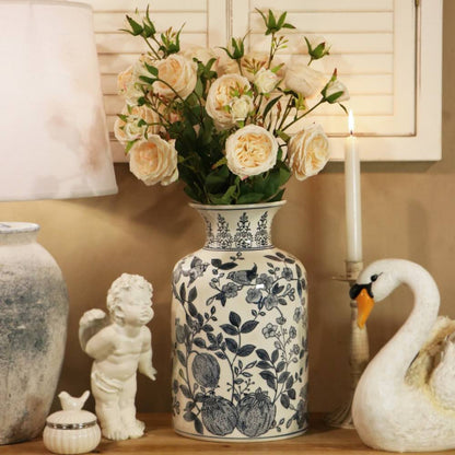 A tall ceramic vase with a blue and white floral pattern, filled with cream-colored faux roses, displayed on a shelf next to a white swan figurine, a small white cherub statue, and a lit candle.