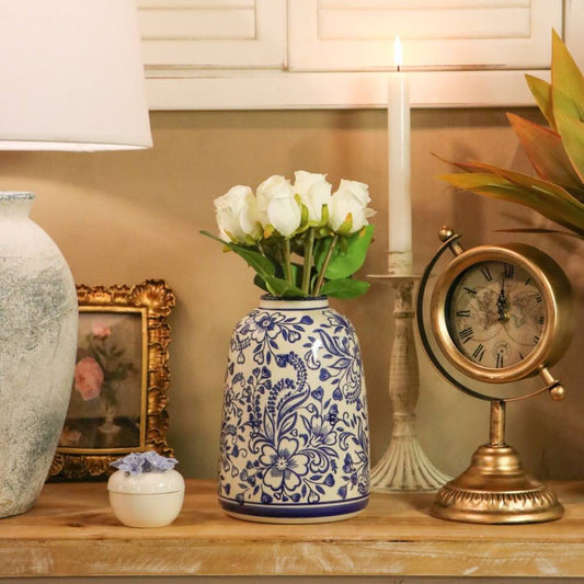 A blue and white floral patterned ceramic vase filled with white roses, styled on a wooden shelf next to a brass desk clock, a candle, and other home decor items.