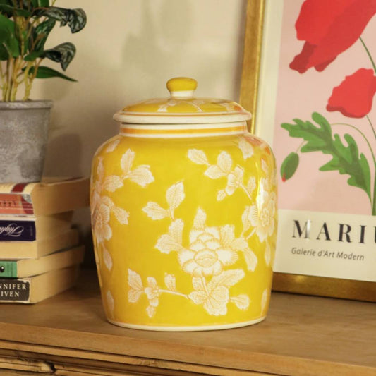 A yellow ceramic lidded jar with a white floral pattern, sitting on a wooden shelf next to books and a framed print.