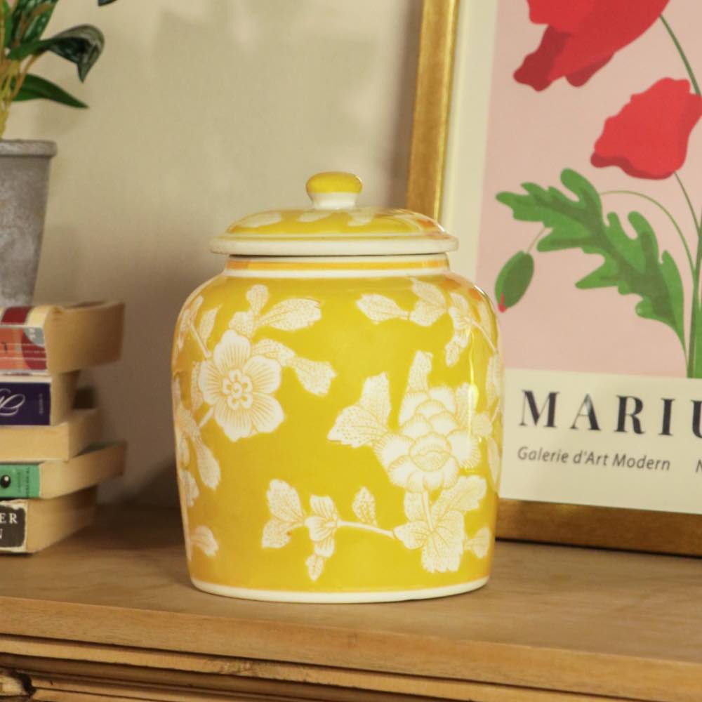 A yellow ceramic jar with a matching lid, decorated with white floral patterns, displayed on a wooden shelf next to a framed print and a small plant.