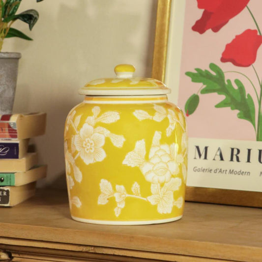 A yellow ceramic jar with a matching lid, decorated with white floral patterns, displayed on a wooden shelf next to a framed print and a small plant.