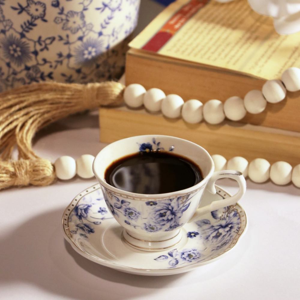 A white ceramic teacup and saucer set with a blue floral pattern, filled with black coffee, styled on a tabletop with books, a tassel, and white beads in the background.