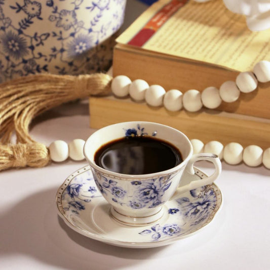 A white ceramic teacup and saucer set with a blue floral pattern, filled with black coffee, styled on a tabletop with books, a tassel, and white beads in the background.