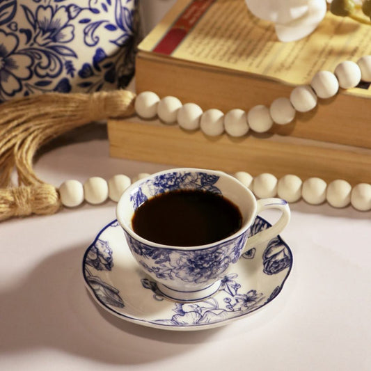 A white ceramic tea cup and saucer with blue floral patterns, filled with black coffee, styled on a tabletop with books, a beaded necklace, and a decorative vase in the background.