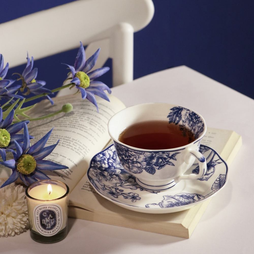 A white ceramic teacup and saucer with a blue floral toile pattern, filled with tea, styled on an open book with blue flowers, a candle, and a dark blue background.