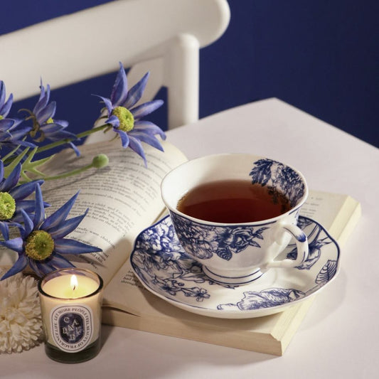 A white ceramic teacup and saucer with a blue floral toile pattern, filled with tea, styled on an open book with blue flowers, a candle, and a dark blue background.