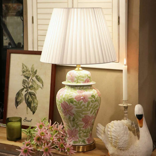 A decorative table lamp with a pleated white shade and a ceramic base featuring a pink and green floral pattern, styled on a tabletop with candles, flowers, and a swan figurine.