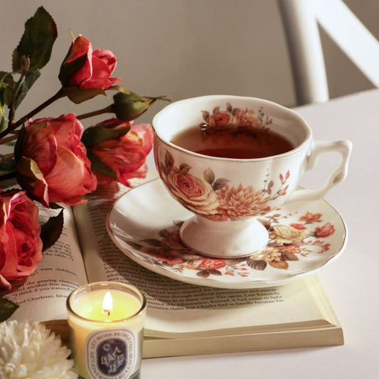 A white ceramic teacup and saucer set decorated with a vintage-style floral print, filled with tea and styled on an open book with red roses and a lit candle.