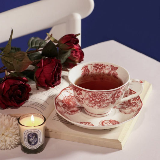 A white ceramic teacup and saucer with a red floral toile pattern, filled with tea, styled on an open book with red roses, a lit candle, and a white chair against a dark blue background.
