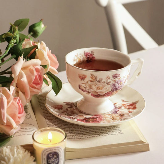 A white porcelain teacup and saucer set with a floral print, filled with tea, styled on an open book with pink roses and a lit candle nearby.