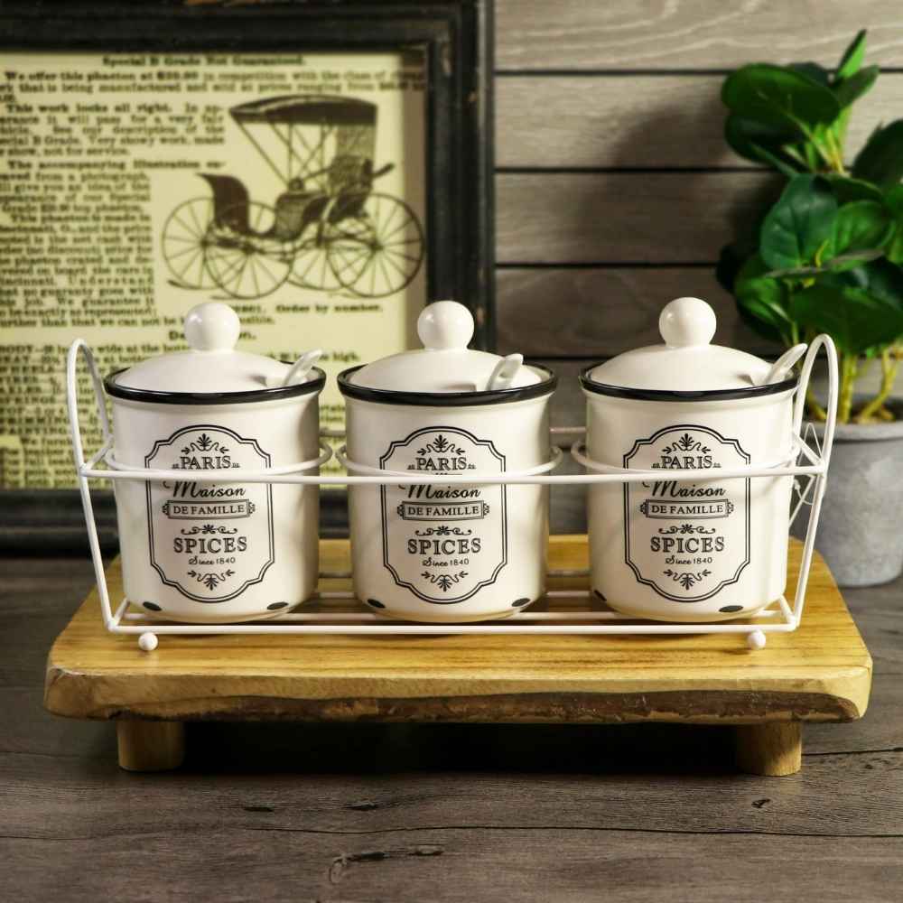 Three white ceramic spice jars with black labels and lids, displayed in a white wire rack on a wooden tray against a rustic background with a framed print and green leaves.