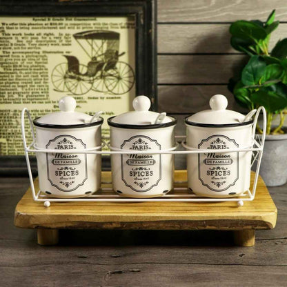 Three white ceramic spice jars with black labels and lids, displayed in a white wire rack on a wooden tray against a rustic background with a framed print and green leaves.