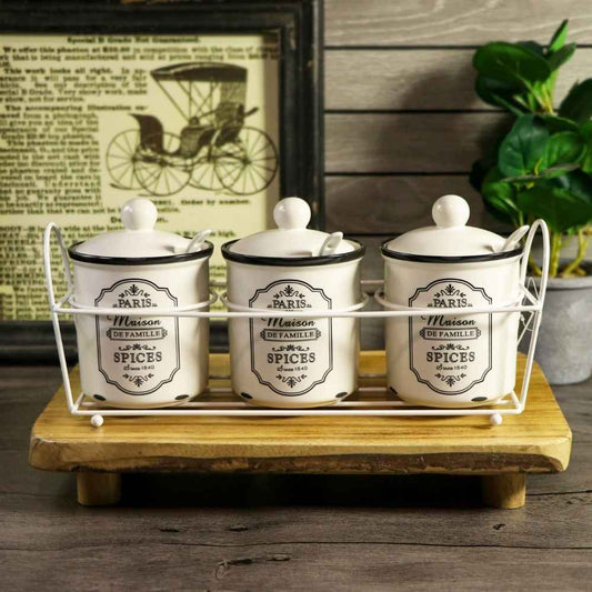 Three white ceramic spice jars with black labels and lids, displayed in a white wire rack on a wooden tray against a rustic background with a framed print and green leaves.