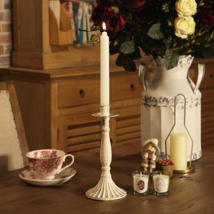 A tall white taper candle in a ribbed cream-colored metal holder sits on a wooden table, styled with a matching teacup, small candles, a decorative figurine, and a large white pitcher filled with red and yellow roses.