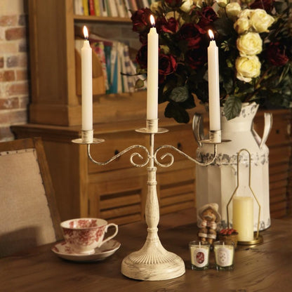 A vintage-style three-arm metal candelabra with an off-white distressed finish stands on a wooden table, holding three lit white taper candles. The scene is styled with a floral arrangement, a teacup, small candles, and a white pitcher in a warm indoor setting.