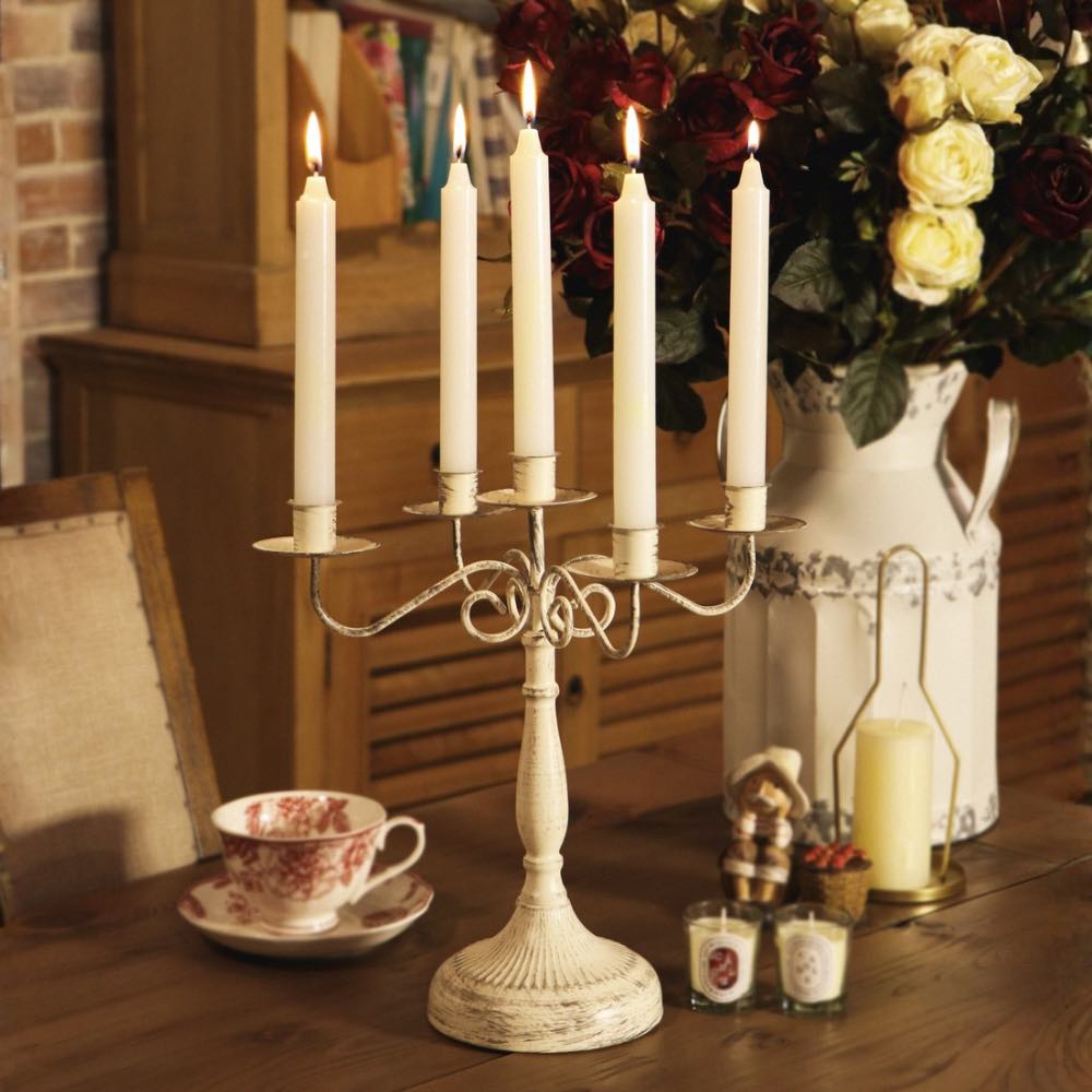 A distressed white five-arm candelabra candle holder with lit taper candles, displayed on a wooden table in a cozy, rustic room setting alongside a floral arrangement, a teacup, and small tea lights.