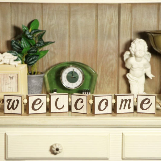 Wooden block letters spelling 'WELCOME' arranged on a tabletop in front of a decorative shelf with a green clock, plant, cherub statue, and other home decor.