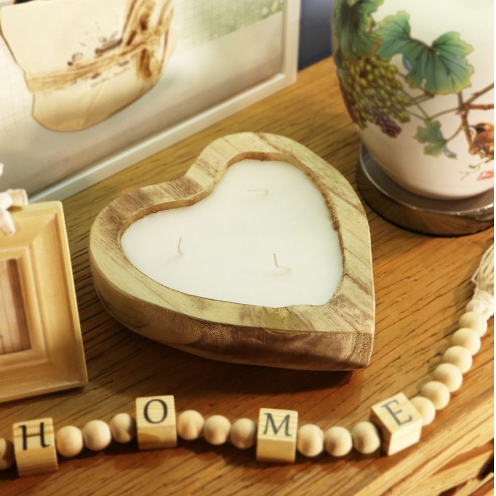 Heart-shaped wooden candle holder with white wax, styled on a wooden table with decorative beads spelling HOME and a ceramic vase in the background.