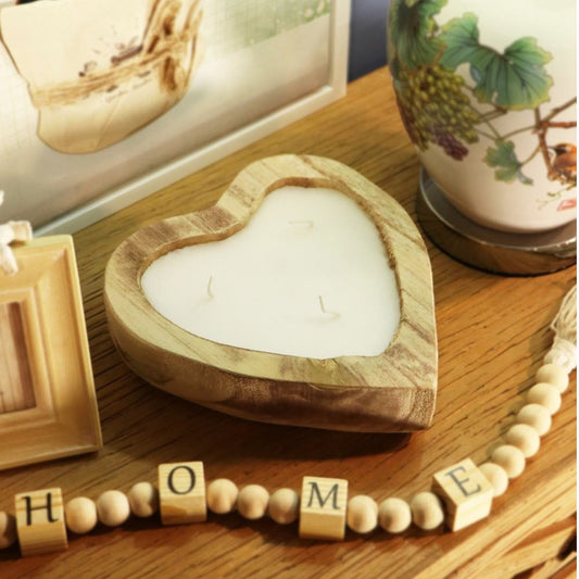 Heart-shaped wooden candle holder with white wax, styled on a wooden table with decorative beads spelling HOME and a ceramic vase in the background.