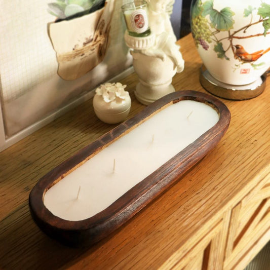 A rectangular wooden tray candle with a dark brown wood frame and white wax, resting on a wooden table next to decorative ceramic items.